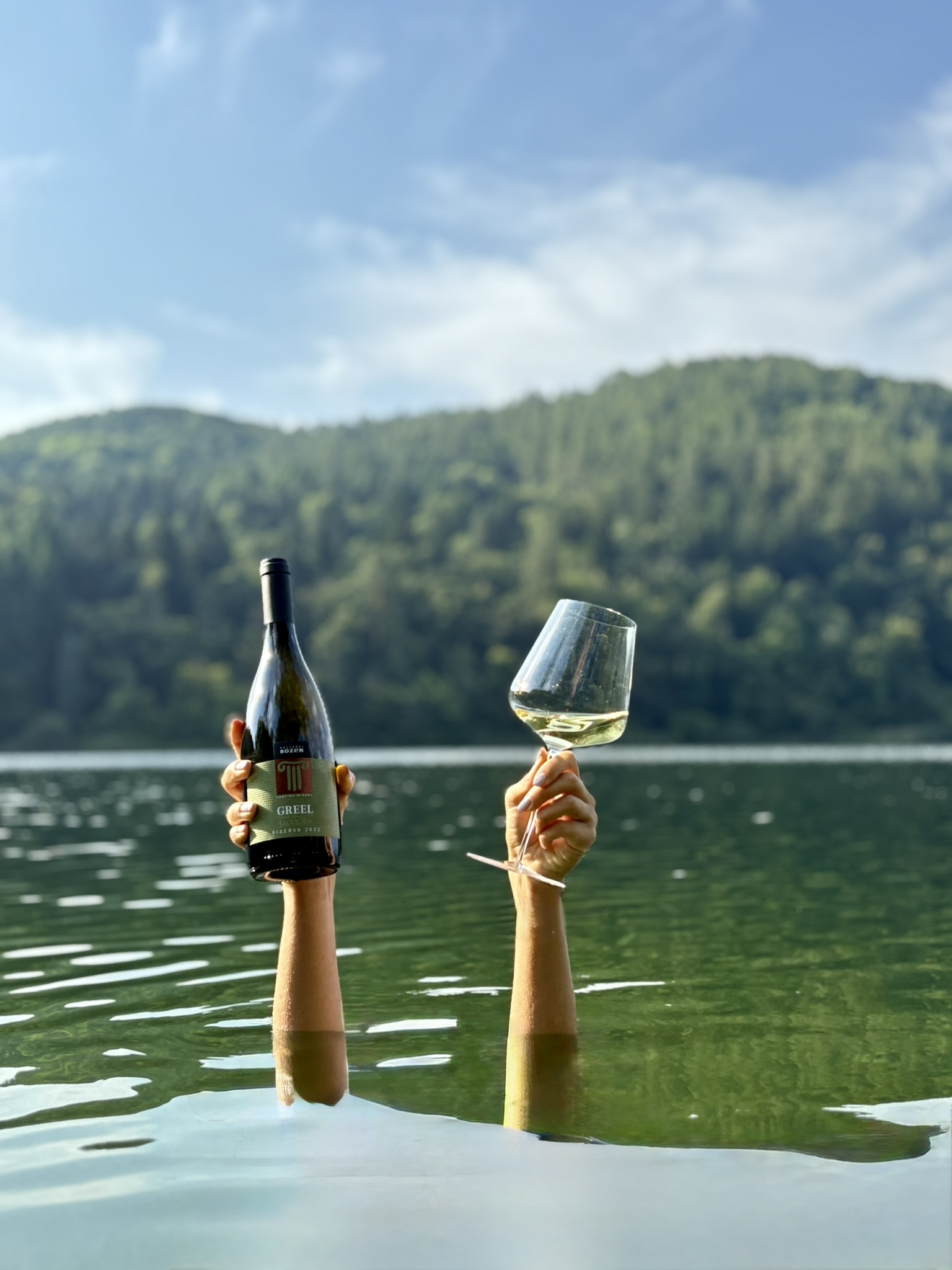 Frau im See untergetaucht hält eine Flasche Kellerei Bozen Greel und ein Glas Weißwein aus dem Wasser